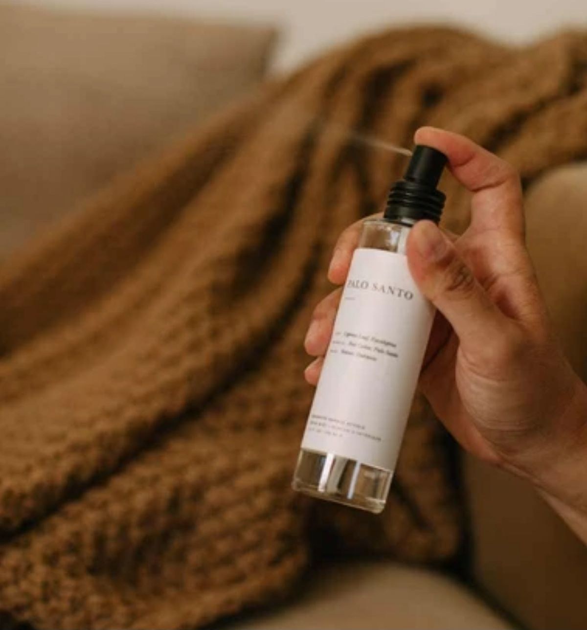 A hand spraying a glass bottle of room mist over a knitted blanket