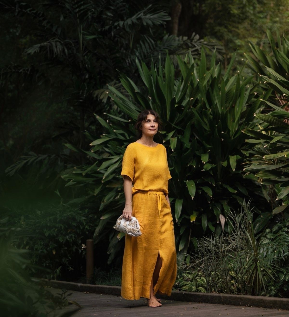 A woman in a mustard linen top and skirt standing on a path in a lush garden