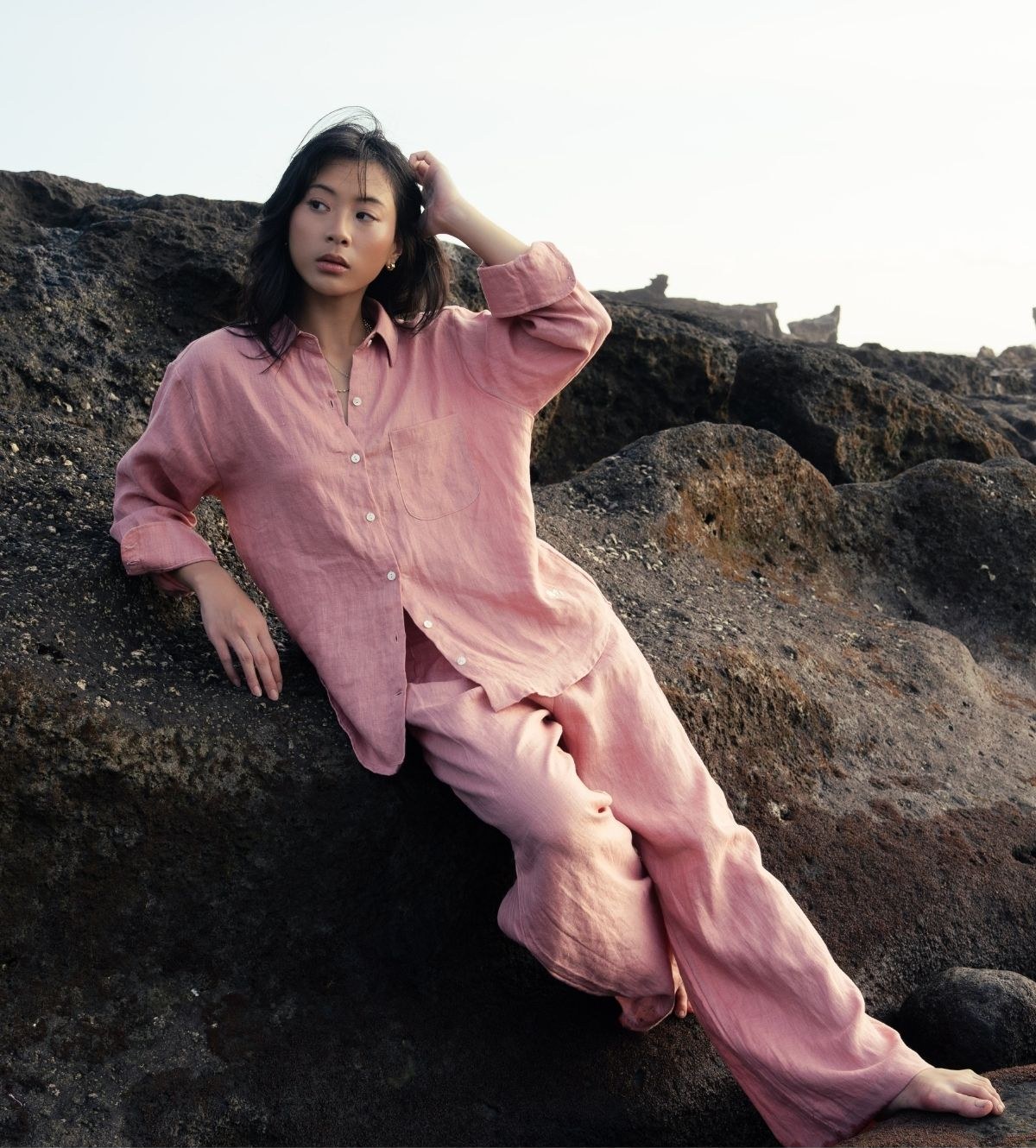 A woman in a pink linen shirt and trousers leaning on dark volcanic rocks