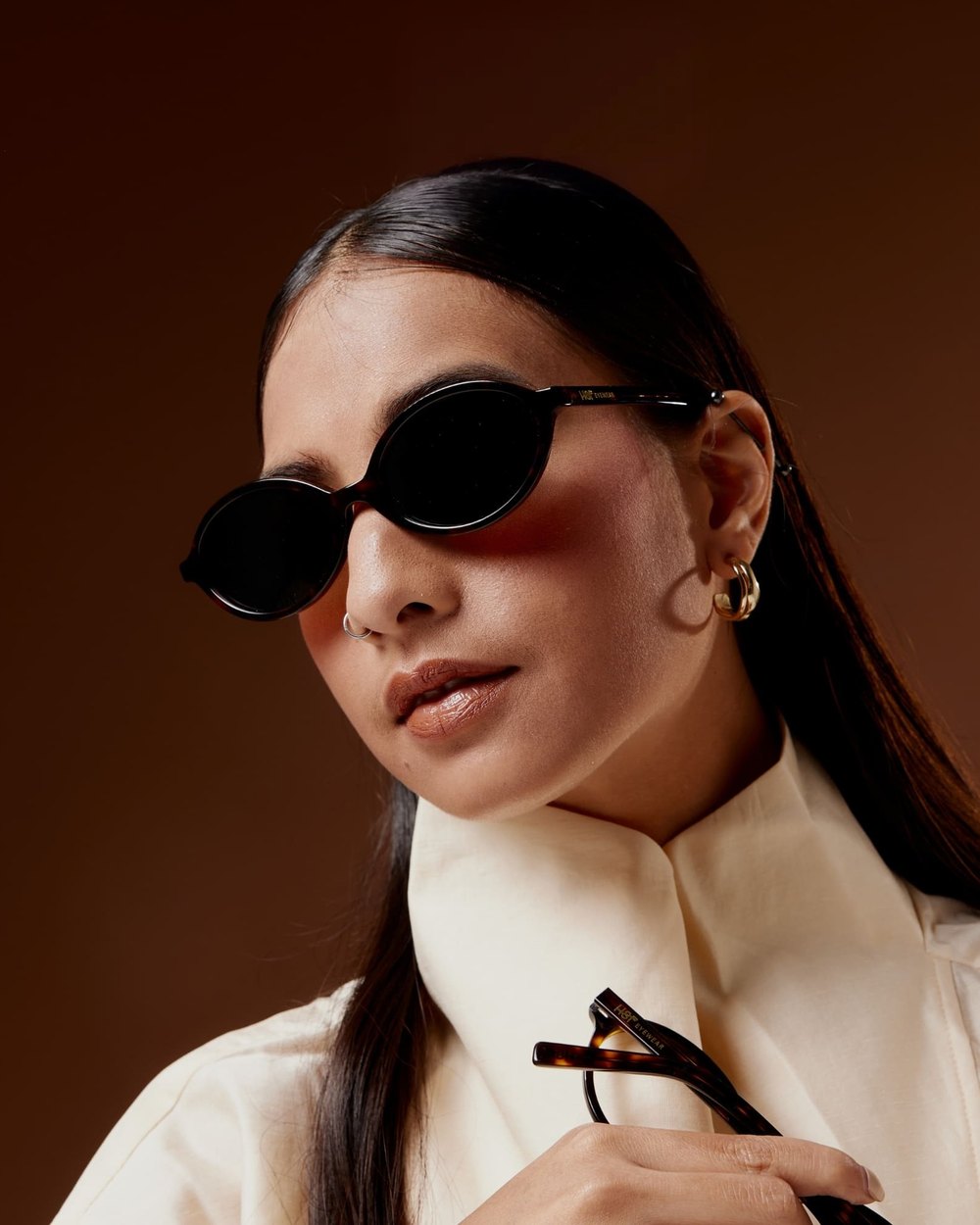 A woman in dark oval tortoiseshell sunglasses against a warm brown backdrop, holding a second pair of frames