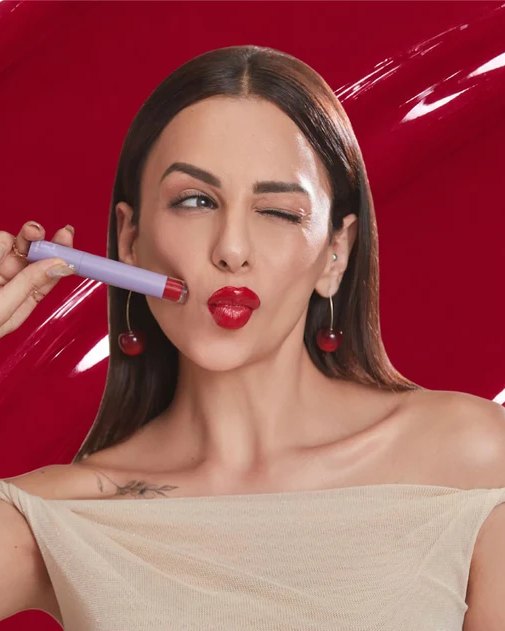 A woman winking and holding a lilac lip product, against a glossy red backdrop