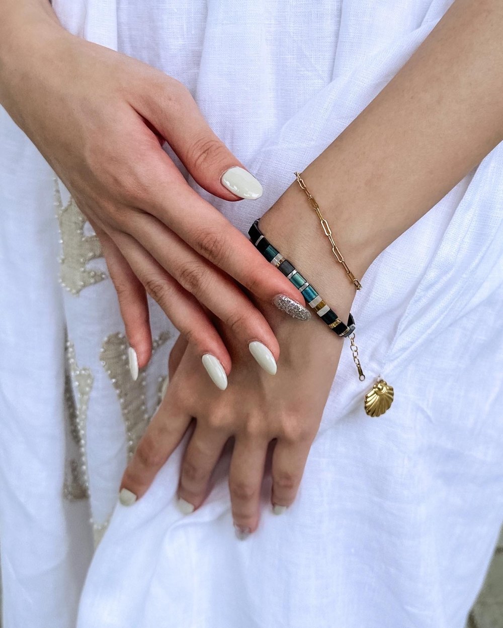 Two hands resting on white linen, wearing a tiled beaded bracelet and a gold paperclip-chain bracelet with a shell charm